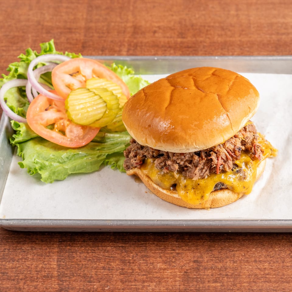 Pulled pork cheeseburger with lettuce, tomato, onion, and pickles on a metal tray.