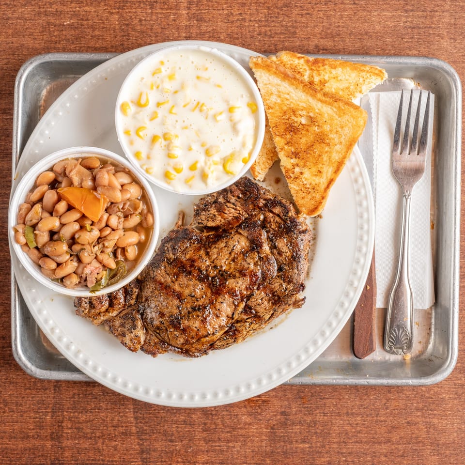 Grilled steak, baked beans, creamed corn, and Texas toast on a white plate with a fork.