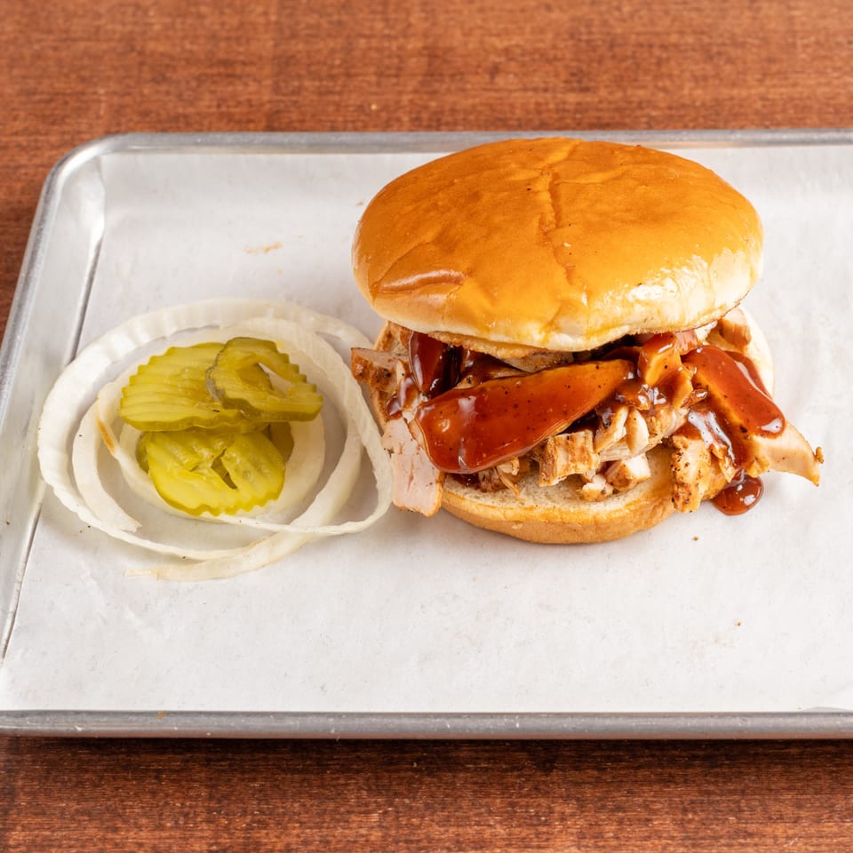 BBQ chicken sandwich on a bun with pickles and sliced onions on parchment paper.